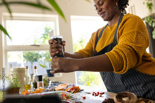 Happy plus size african american woman in apron seasoning vegetables in kitchen