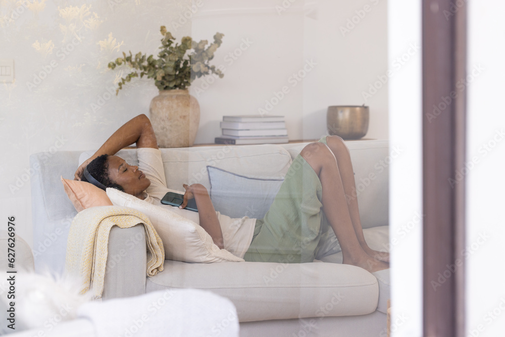 Relaxed african american woman wearing headphones lying on sofa with smartphone