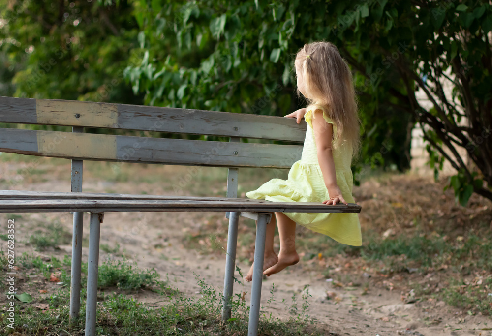 Naklejka premium lonely little child girl from the back in yellow dress sitting on a bench looking at the distance outdoors in nature