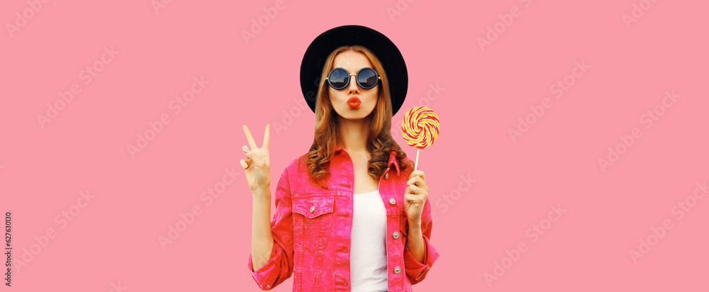 Portrait of happy young woman holding lollipop and blowing her lips sends air kiss wearing pink jacket, black round hat on pink background