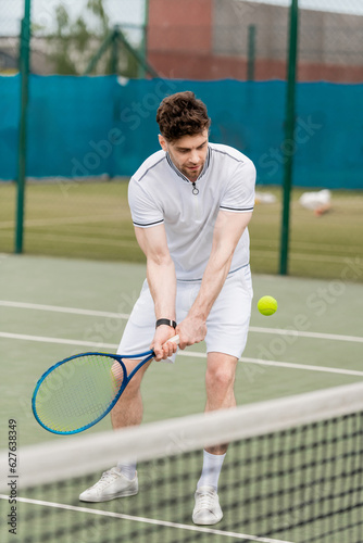 handsome tennis player holding racket and hitting tennis ball on court, sport as a hobby