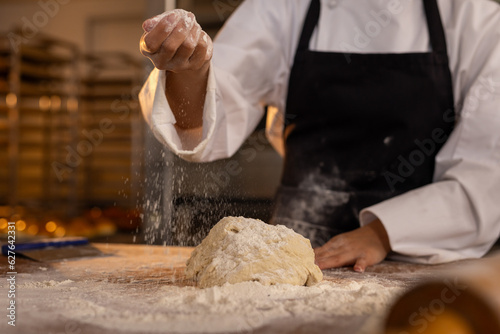 Asian female baker wearing apron in bakery kitchen and pouring flour on dough photo