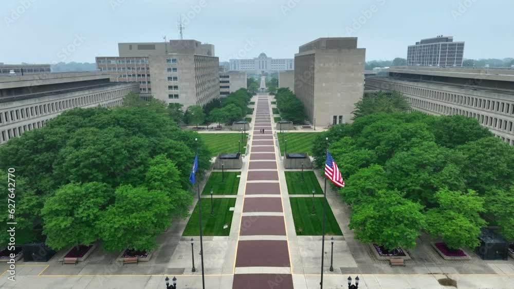 American flag and Michigan state flag waving in government complex with many offices for government employees. Aerial establishing shot of Lansing, MI capitol building grounds.