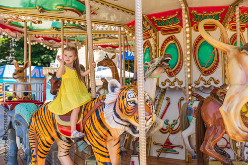 Adorable little blonde girl in summer yellow dress at amusement park having a ride on the merry-go-round. Child girl has fun outdoor on sunny summer day. Entertainment concept