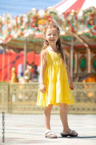 Adorable little blonde girl in summer yellow dress at amusement park. Cute girl stands on the background of the carousel and laughs. Entertainment concept