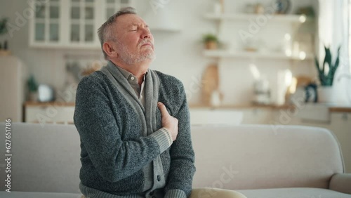 Senior man suffers from heartache sitting on couch at home. Unwell old male with chest pain myocardial infarction has chronic cardiology disease. Cardiac arrhythmia, heart attack, illness concept. photo