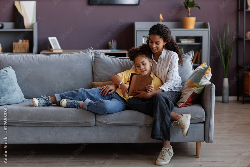 Obraz premium Full length portrait of loving mother and daughter using digital tablet together while relaxing on sofa at home, copy space