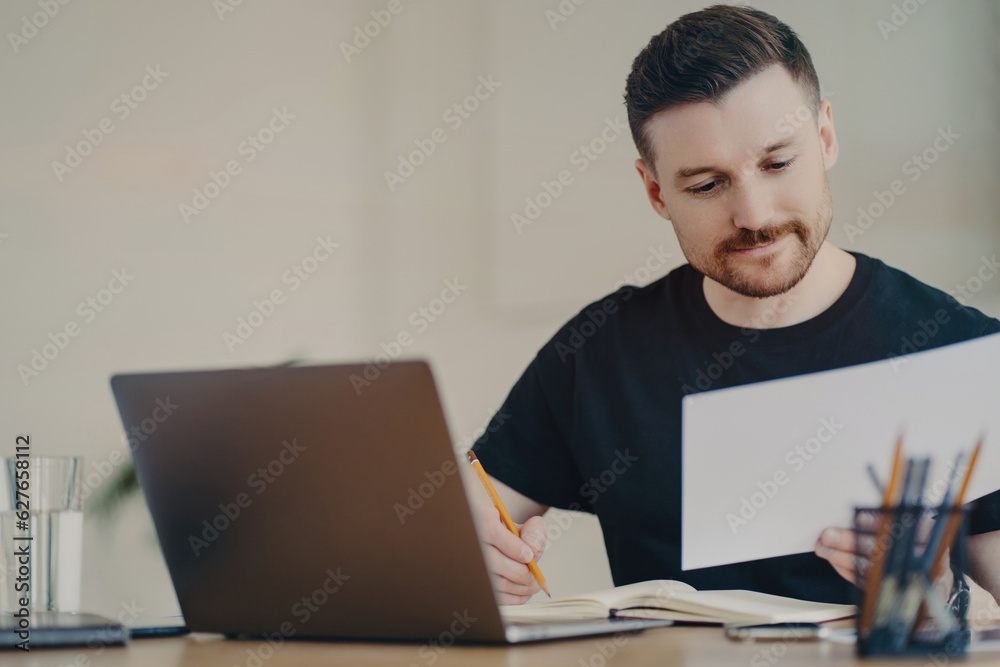 Bearded expert edits reports on laptop, focused at home office, casual in black t-shirt. Bearded expert edits reports on laptop, focused at home office, casual in black t-shirt.