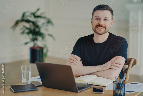 Happy freelancer/businessman, arms crossed, beaming at camera, working remotely from home with laptop and documents.