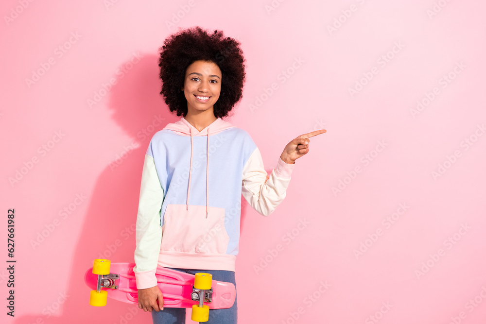 Portrait of cheerful small schoolgirl hand hold skateboard indicate finger empty space promo isolated on pink color background