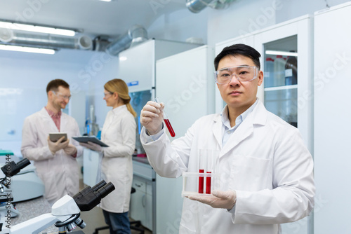 Portrait of a young Asian male doctor  lab technician standing in a lab coat in the clinic and holding tubes with blood  researching for tests  dna. Colleagues are talking in the background.