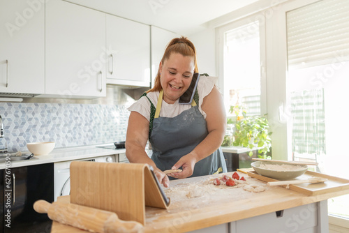 curvy woman smiling in the kitchen preparing a recipe while talking on the phone and watching a tutorial on her tablet