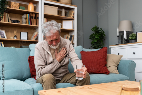 Senior older man getting chest pain in living room, heart attack holding his hand on the chest sitting on sofa at home. Health care older people issue trying to drink water from glass