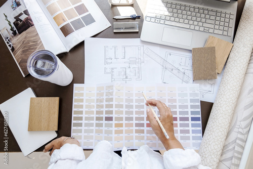 Top view of interior designer or architect in casual wear working on a blueprint for new project, standing in her office on a daytime. Laptop, color samples, coffee cup, tools