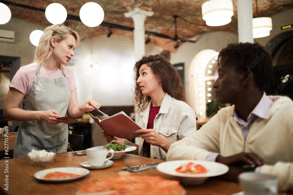 Disgruntled curly-haired young female restaurant guest sitting at table with boyfriend and reading menu while talking to waitress