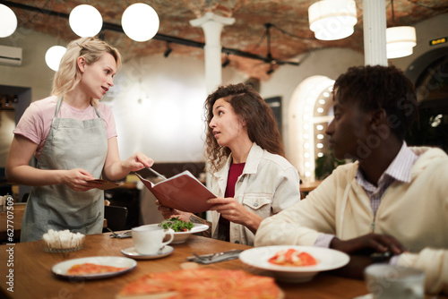 Disgruntled curly-haired young female restaurant guest sitting at table with boyfriend and reading menu while talking to waitress