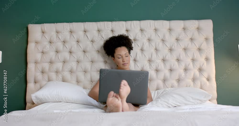 Biracial woman sitting on bed and using laptop in bedroom, slow motion