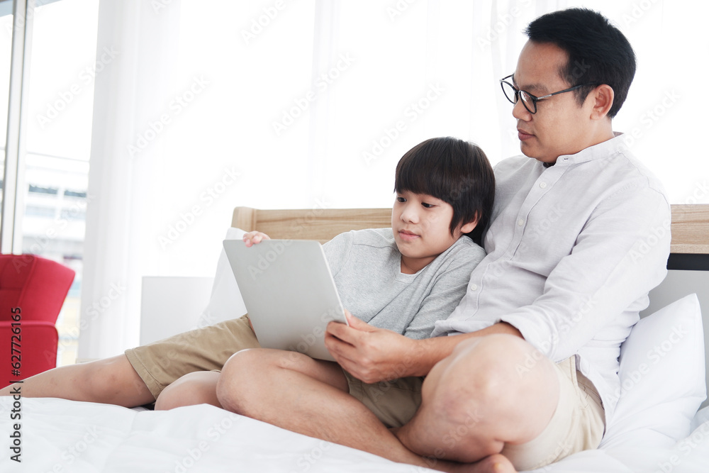 Smiling Asian father and son child is online learning with laptop together on bed in bedroom. Family and education on holiday concept.
