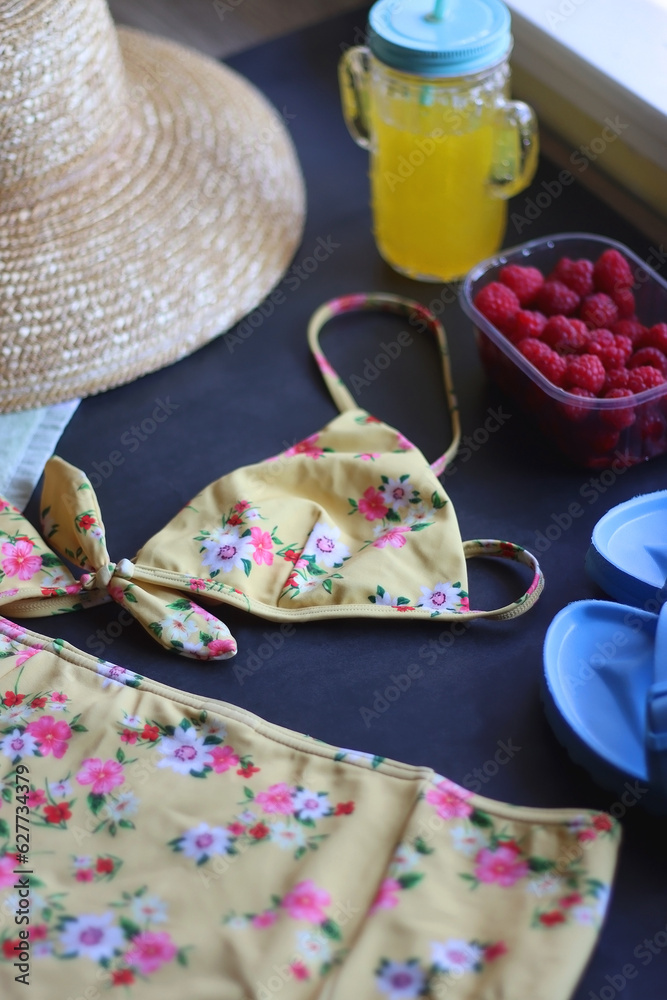 Floral swimsuit, striped towel, straw hat, cactus cup with orange soda, bowl of raspberries and blue sandals. Summer or beach essentials. Selective focus, dark background. Floral swimsuit, striped towel, straw hat, cactus cup with orange soda, bowl of raspberries and blue sandals. Summer or beach essentials. Selective focus, dark background.