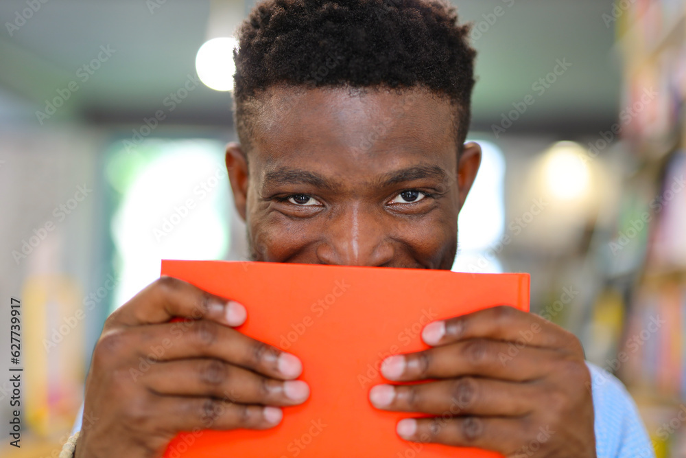 African man holding a bright orange book with both hands in a library setting. The background consists of bookshelves filled with colorful books and is slightly blurred. African man holding a bright orange book with both hands in a library setting. The background consists of bookshelves filled with colorful books and is slightly blurred.