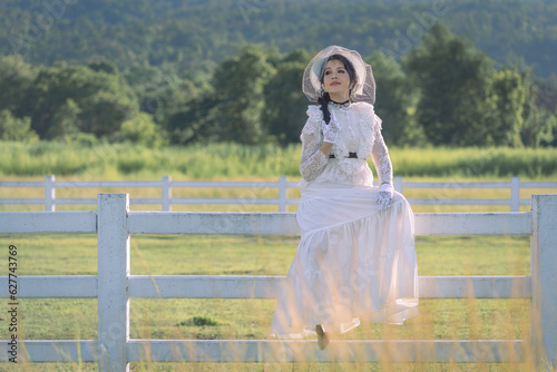 Pretty Asian women in ancient-style costumes sitting on white fence of the stables relax happily together. Vintage blouse costume. Historical dresses, Victorian or Edwardian era style dress.