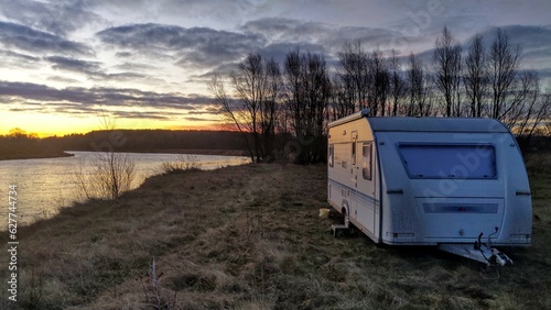 On a grassy riverbank next to trees stands a caravan trailer for a comfortable fishing vacation. The late autumn sun rises and paints the sky and clouds in bright colors There are ripples on the water