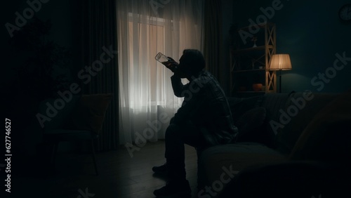 A devastated young man sits in a dark room with a bottle of alcohol, drinking from his throat. Alcoholism. Hopelessness. Alcohol addiction treatment. The concept of mental health.