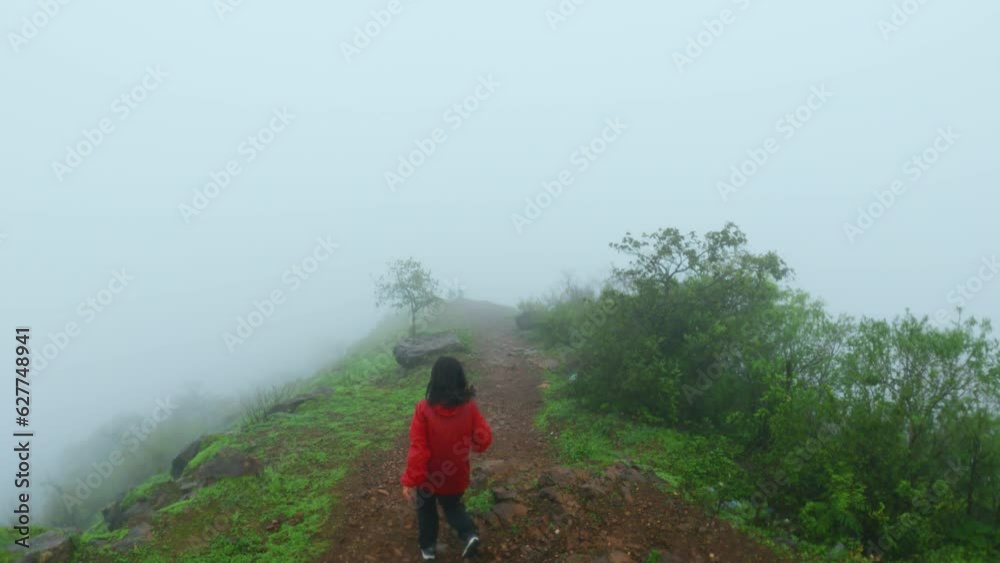 4K shot of Young Indian woman wearing red rain jacket and running with spreading arms on green mountain covered with clouds. Female traveler enjoying cloudy rainy weather. Monsoon in mountains. 4K shot of Young Indian woman wearing red rain jacket and running with spreading arms on green mountain covered with clouds. Female traveler enjoying cloudy rainy weather. Monsoon in mountains.