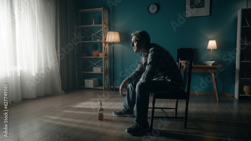 A man sits in a dark room on a chair in front of a window from which light streams. The man looks into the light, holds a glass of alcohol in his hand, there is an almost empty bottle on the floor.
