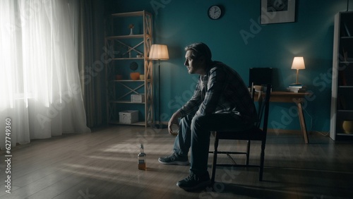 A man sits in a dark room on a chair in front of a window from which light streams. The man looks into the light, holds a glass of alcohol in his hand, there is an almost empty bottle on the floor. photo