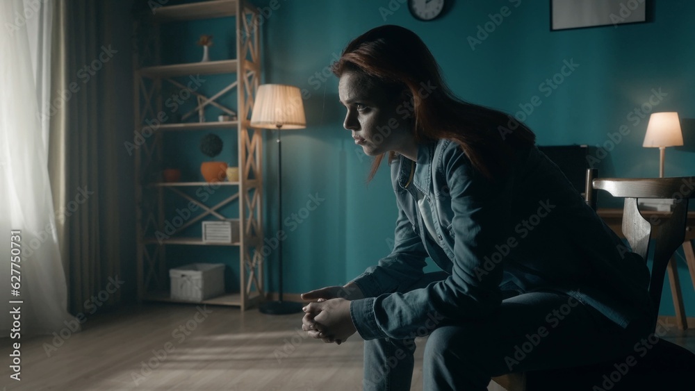 A woman sits on a chair in the middle of the room and looks at the light in the window. Lonely woman sitting in a dark room close up. Concept of hope, mental health.