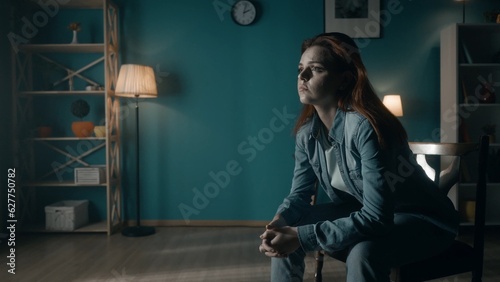 A woman sits on a chair in the middle of the room and looks at the light in the window. Lonely woman sitting in a dark room close up. Concept of hope, mental health.