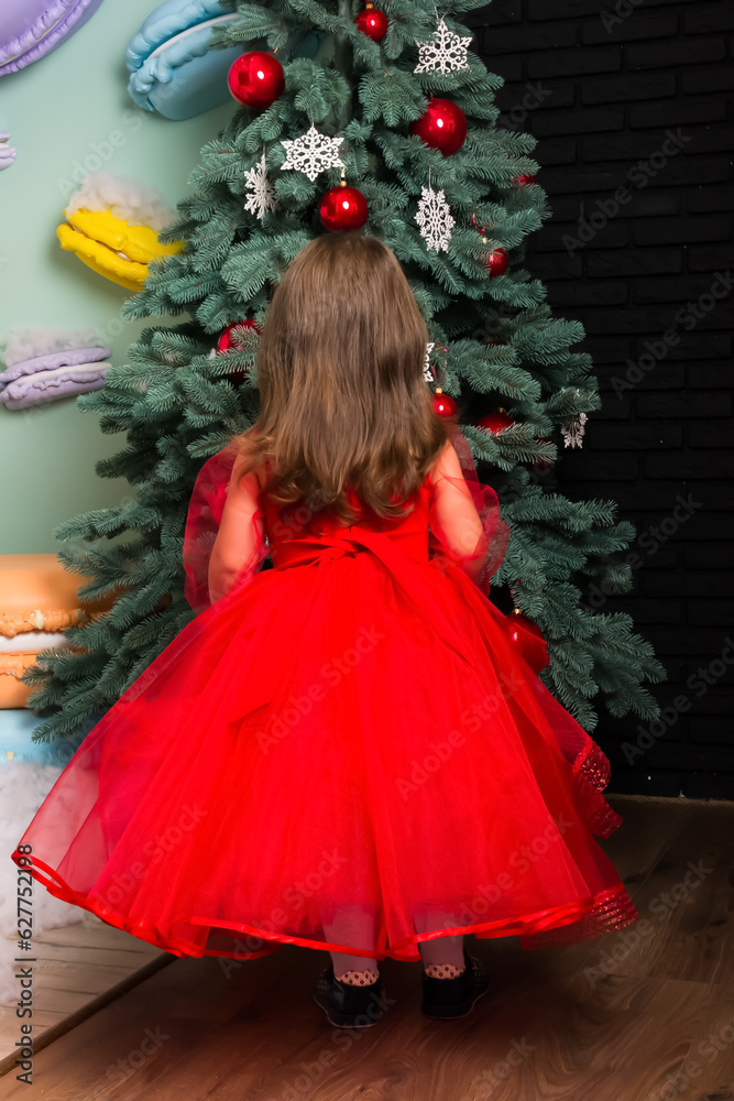 Little girl in red dress stands with his back and decorates the Christmas tree. Little beautiful girl in a red evening dress stands with her back by the Christmas tree. Girl in red in Christmas Little girl in red dress stands with his back and decorates the Christmas tree. Little beautiful girl in a red evening dress stands with her back by the Christmas tree. Girl in red in Christmas