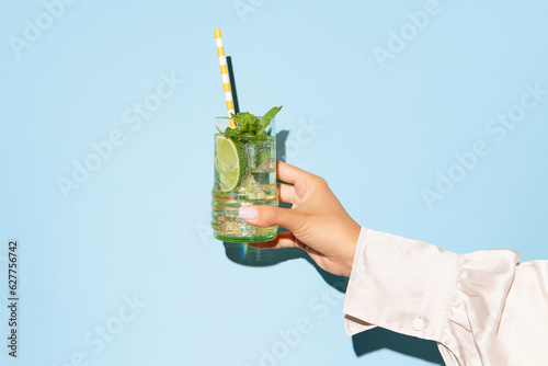 Lady's hand holding glass of mojito with lime on light blue background.