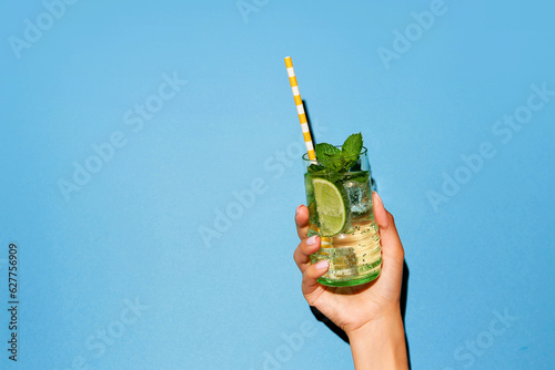 Lady's hand holding glass of mojito with lime and bright straw on light blue background.