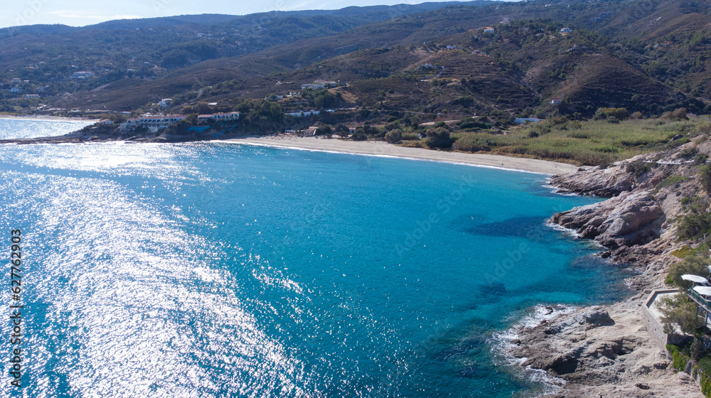 The Livadi Beach at the Ikaria island in a quiet summer day with blue clear water and nature behind, Ikaria, Greece. Little beach lagoon next to the shore.