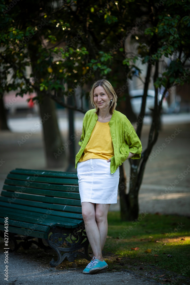 A woman wearing a white skirt and a lettuce-colored jacket stands in a city square.