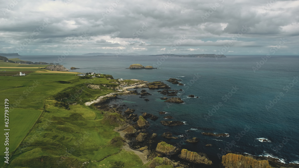 Rocky shore aerial view: green grass meadows and fields. Water surface stretches to horizon with grey clouds. Alone building on cliff. Northern Ireland panoramic landscape.