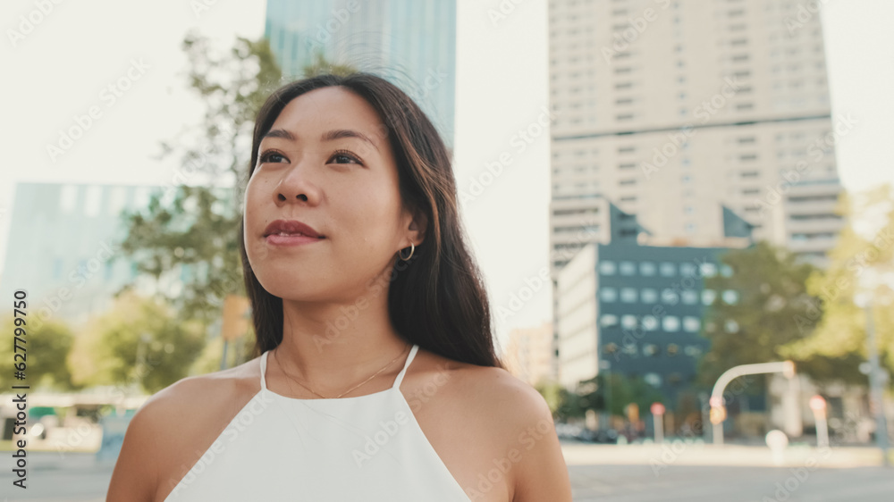 Close-up of lovely brunette girl dressed in white top and jeans, looking around at modern city background