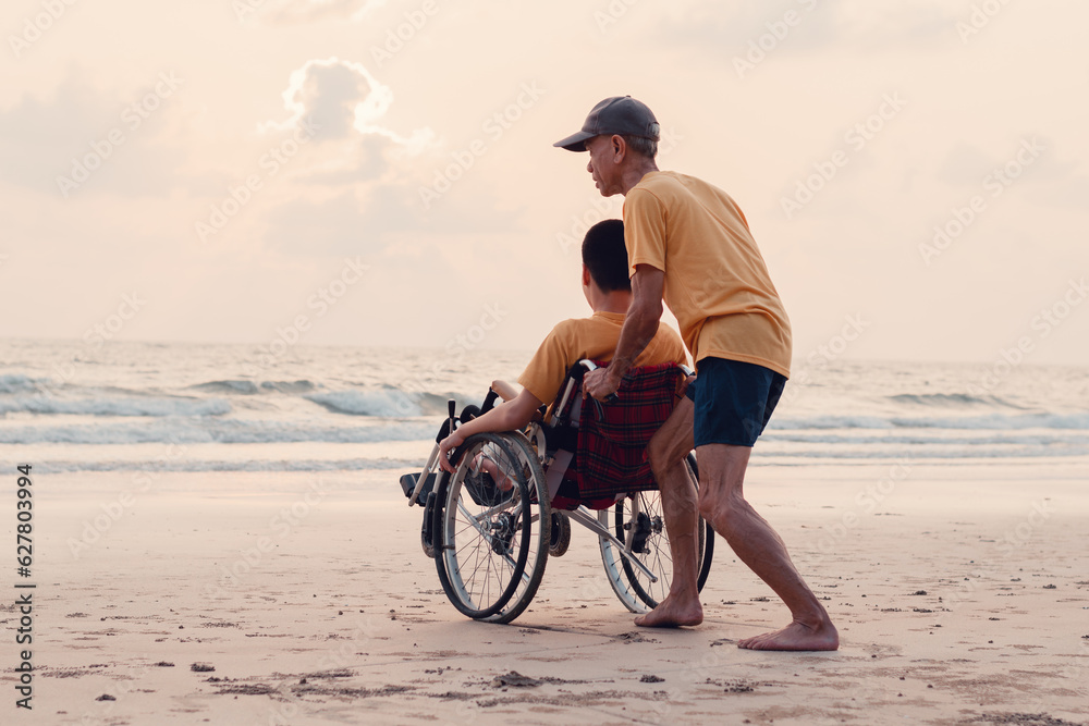 Fototapeta premium A parent, caregiver or volunteer helping teenage boy push wheelchair to the beach in evening sunset,exercise and learning about nature on the sea,Travel on vacation with family, Mental health concept.