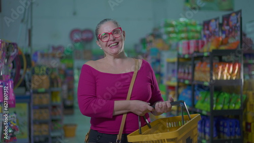 Happy female shopper holding basket standing inside supermarket. Middle-aged woman smiling at grocery store depicting consumerism lifestyle habits