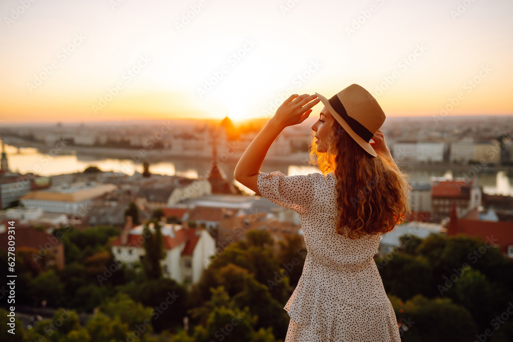 Fototapeta premium Happy traveler in a stylish dress and hat enjoys the sunrise or sunset with stunning views of the city. Back view. Lifestyle, travel, tourism, nature, active lifestyle.