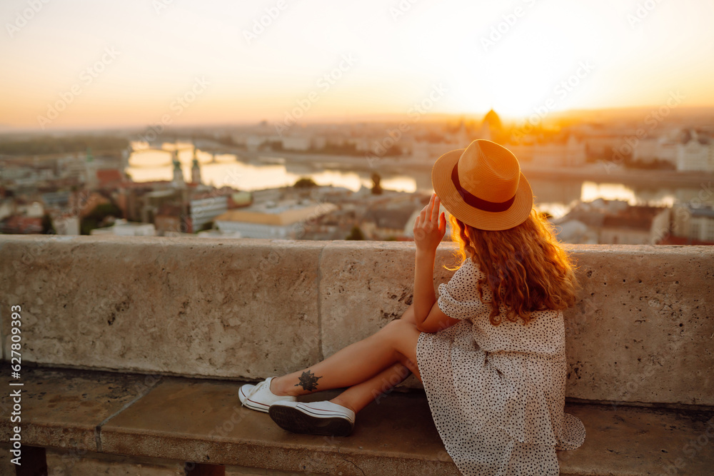 Happy traveler in a stylish dress and hat enjoys the sunrise or sunset with stunning views of the city. Back view. Lifestyle, travel, tourism, nature, active lifestyle.