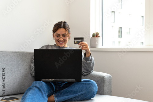 young black-haired woman shopping online from the sofa at home with the computer and credit or debit card