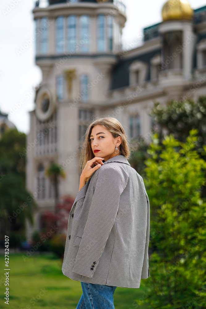 Woman standing on street. Beautiful young woman wearing jacket and jeans walking. Green trees and historic building facade on background. City recreation. Carefree person resting experiencing calmness Woman standing on street. Beautiful young woman wearing jacket and jeans walking. Green trees and historic building facade on background. City recreation. Carefree person resting experiencing calmness