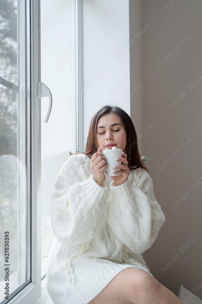 Woman in warm white winter sweater standing next to the window at home at christmas eve holding cup with marshmallows, fir tree behind Woman in warm white winter sweater standing next to the window at home at christmas eve holding cup with marshmallows, fir tree behind