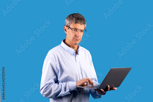 Mature businessman holding laptop frowning typing on blue studio background photo