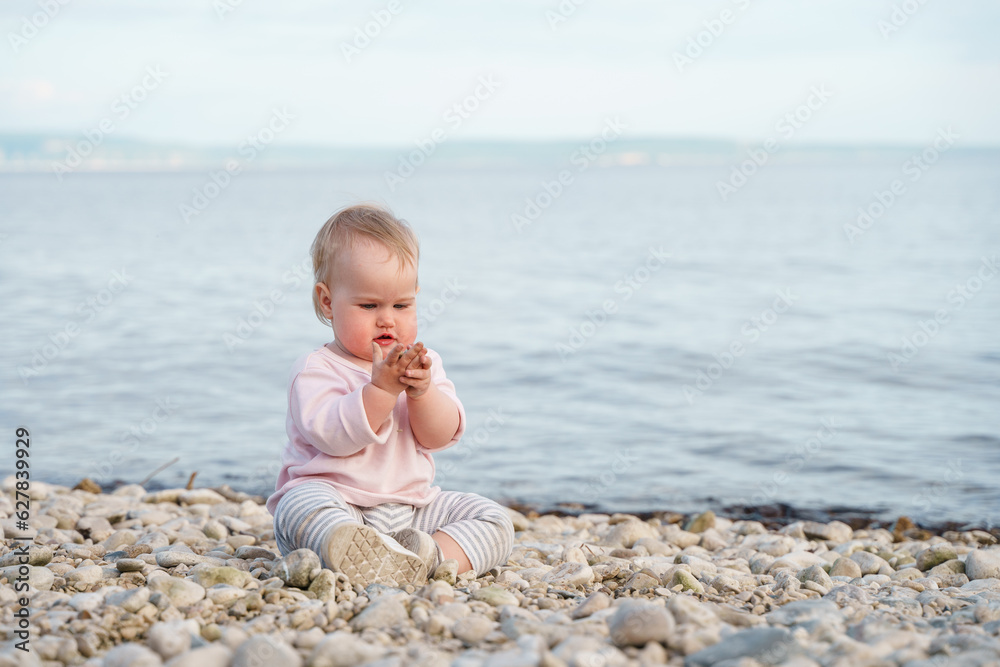A one-year-old toddler sits on the beach of the sea in the evening after sunset A one-year-old toddler sits on the beach of the sea in the evening after sunset