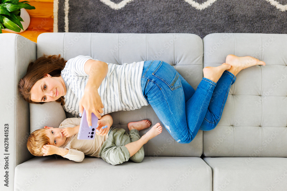 Woman and her little son lying on cozy sofa with smartphone, above view Woman and her little son lying on cozy sofa with smartphone, above view