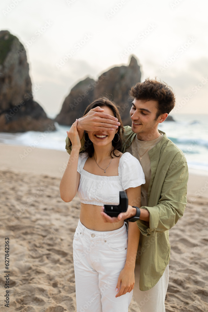 Loving european guy making proposal to his lady, closing her eyes and holding gift box with ring on beach, vertical shot